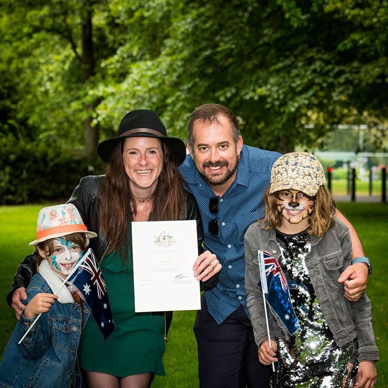 Happy family at citizenship ceremony, gathered around certificate, 2 kids wearing animal face paint, holding Australian flags