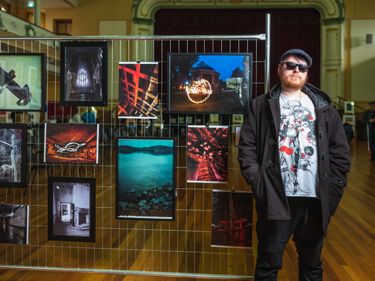A man with glasses and a cap standing next to his photography exhibit at City Hall