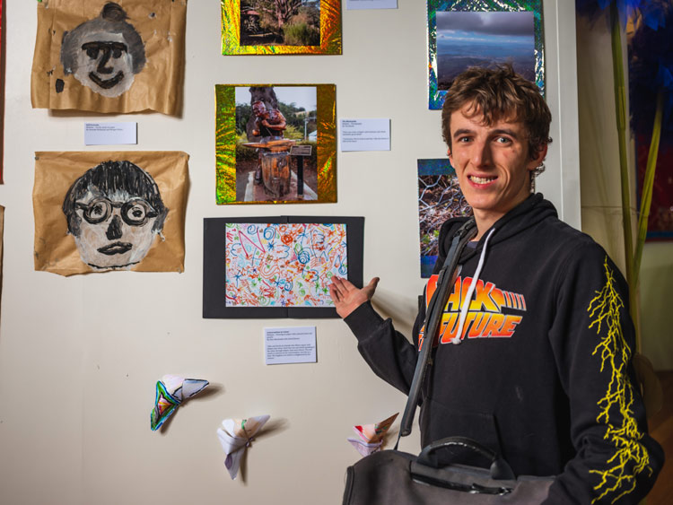 A young man standing in front of an art display pointing to his drawing