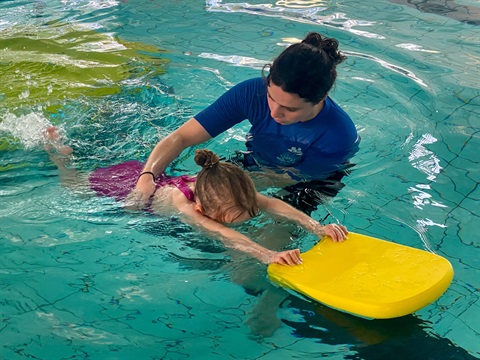 A hobart learn to swim instructor in blue rashie is guiding a young blind girl through the water while holding a kickboard 