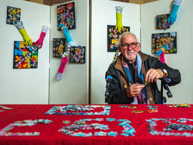 A man sitting at a table completing jigsaw puzzles with a display of framed art work behind him