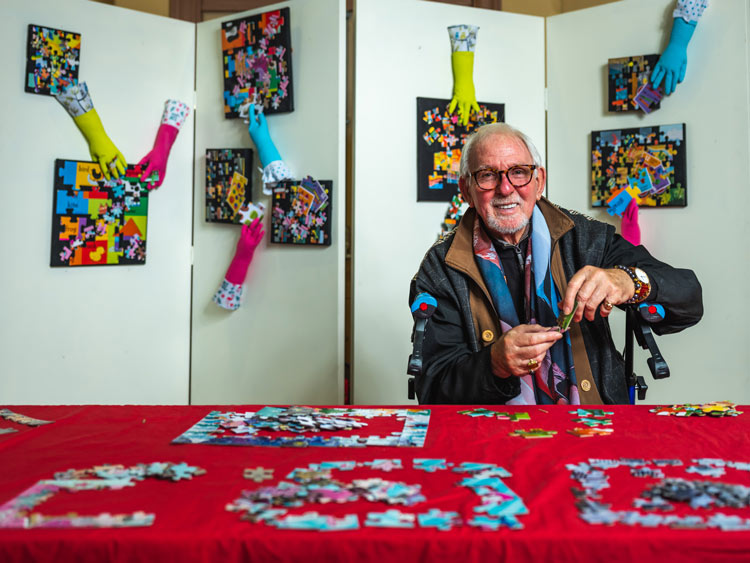 A man sitting at a table completing jigsaw puzzles with a display of framed art work behind him A man sitting at a table completing jigsaw puzzles with a display of framed art work behind him