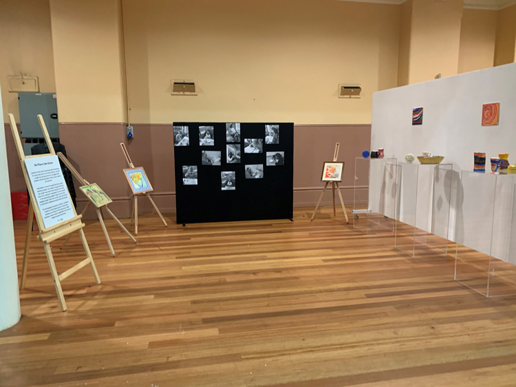 Black and white photographs hanging on a black screen, paintings on easels along with pottery displayed on clear plinths by Mosaic Support Services