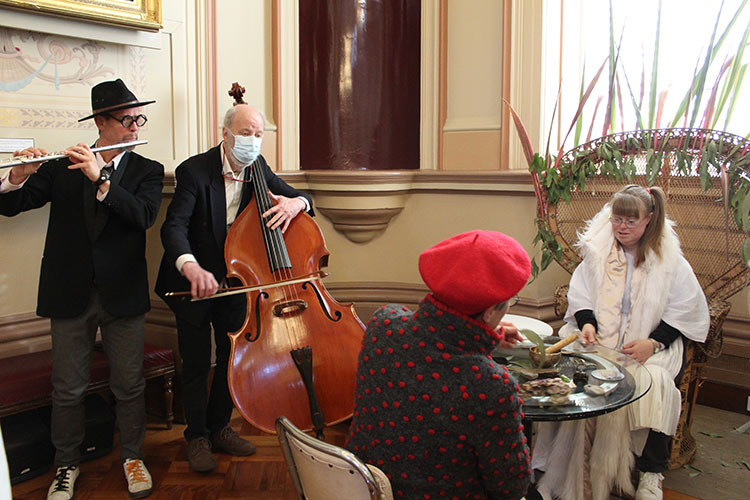 Two people sitting at a table with two musicians standing next to them playing cello and flute.