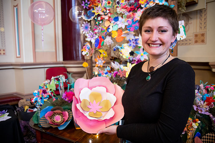 Artist holding a large paper flower with the community flower installation behind.