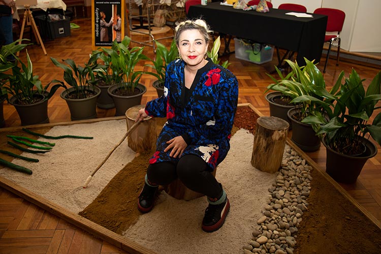 Artist sitting on a wooden stump in a sandpit with coloured sand and rocks, holding a stick.